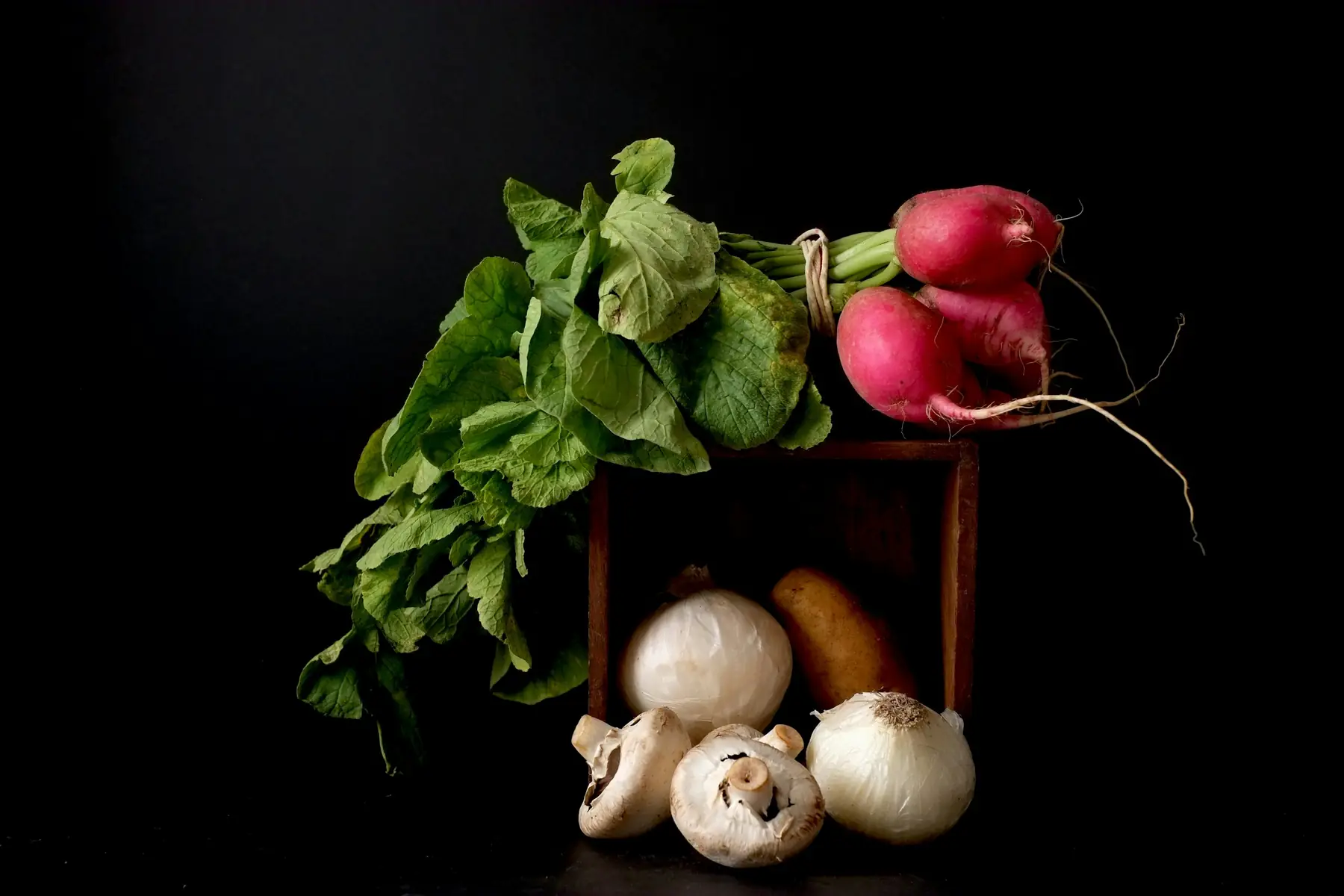 Leaves, radishes, mushrooms and onions in a wooden crate, lit against a dark background